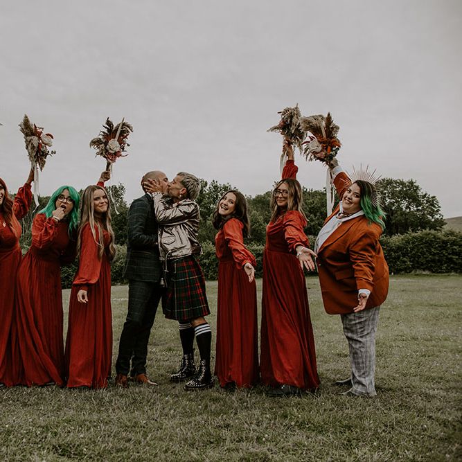 Grooms kiss in the middle of their bridesmaids who wear deep red gowns from ASOS and hold homemade floral bouquets 