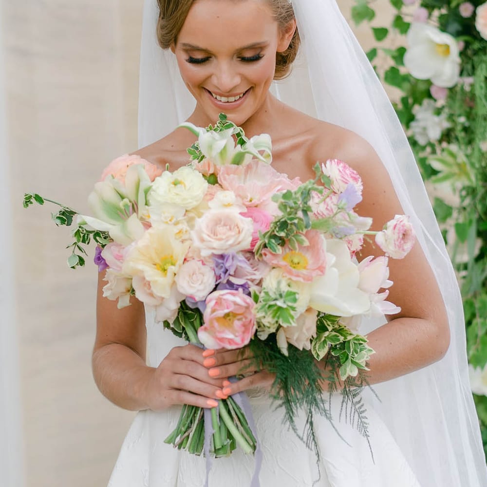 Bride wearing a beaded pearl headband smiles as she looks at her pretty bridal bouquet of pink, white and purple flowers with roses and lilies 