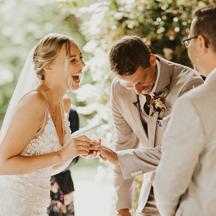 Bride laughs wearing a lace wedding dress and veil as he she puts on the groom's wedding band during the ceremony 