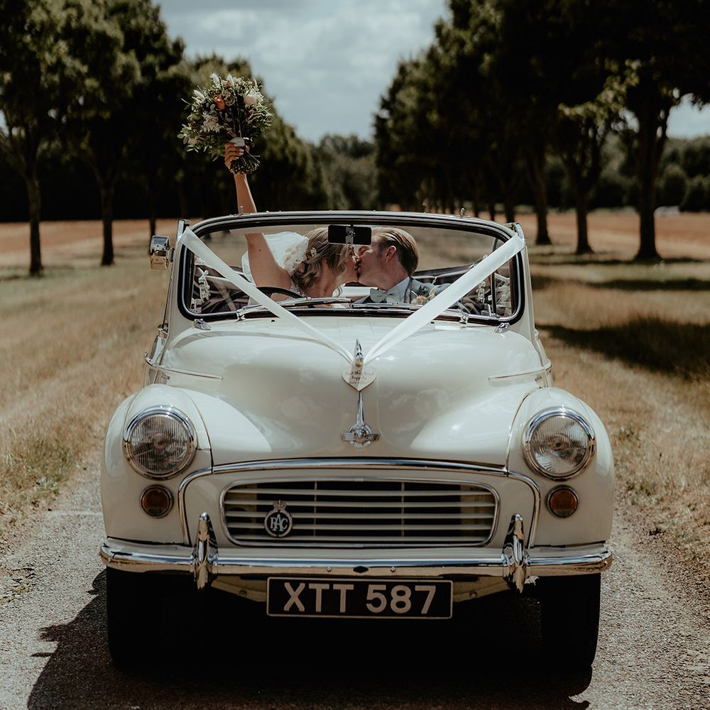 Cream vintage convertible wedding car with white ribbon decorations and flowers with the bride and groom riding inside 
