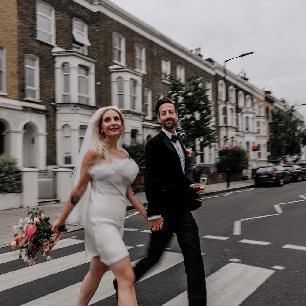 Bride in white short satin dress with feather trimmed neckline and groom in black tux with bowtie walking across the road 