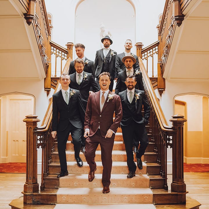 Groom in maroon suit walks down staircase with his groomsmen in blue suits 