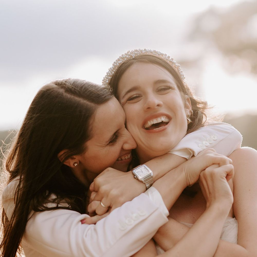The bride in a white suit jacket and silver watch hugs the bride in a pearl headband and off the shoulder wedding dress from behind 