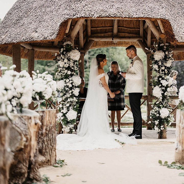 The groom reads out his vows to the bride as they stand next to white flower columns for their outdoor wedding ceremony  