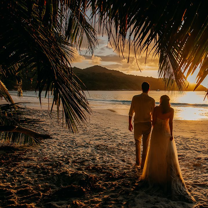 Bride in off the shoulder lace wedding dress and groom in white shirt and chinos walking on the beach in Seychelles at sunset 