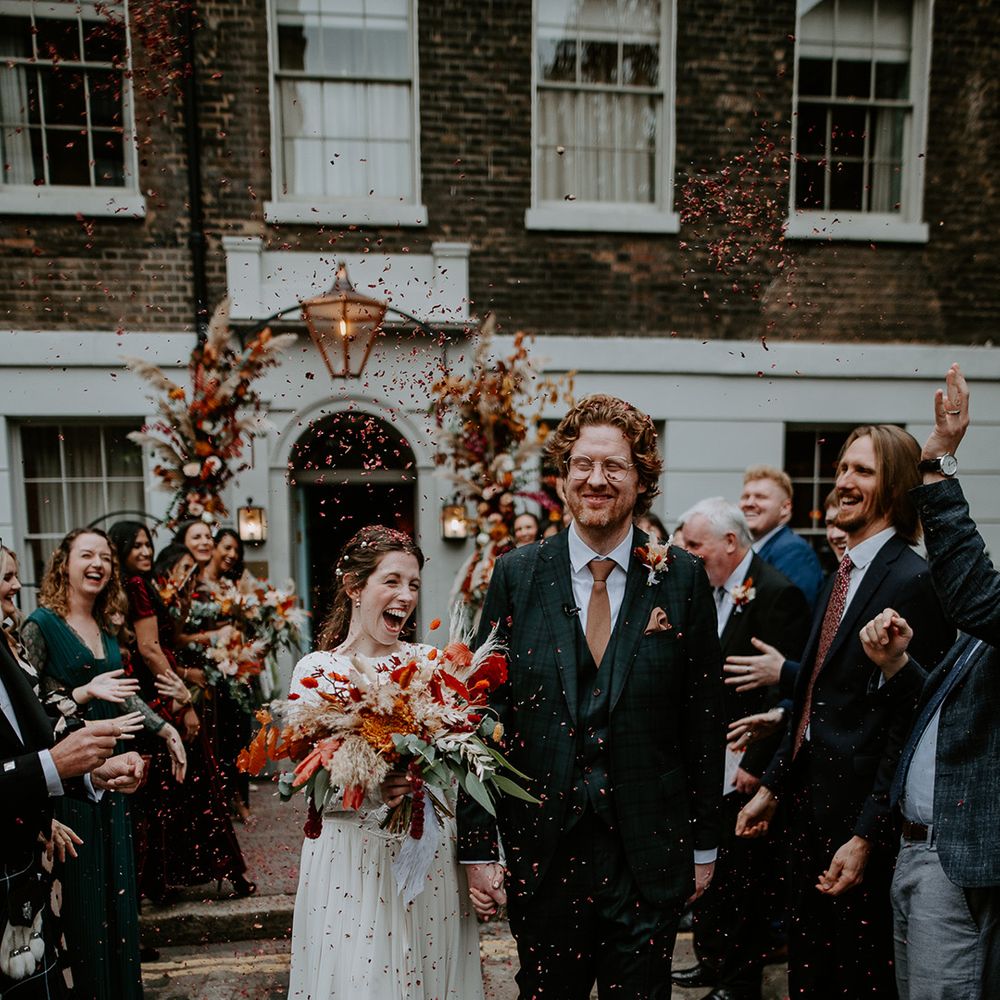 Bride in long sleeve lace vintage wedding dress and closed toe satin heels holding large autumnal bouquet with orange, black, green and yellow roses, peonies, foliage and pampas grass with groom in dark suit with orange tie and autumnal boutonniere doing confetti walk outside The Zetter Townhouse