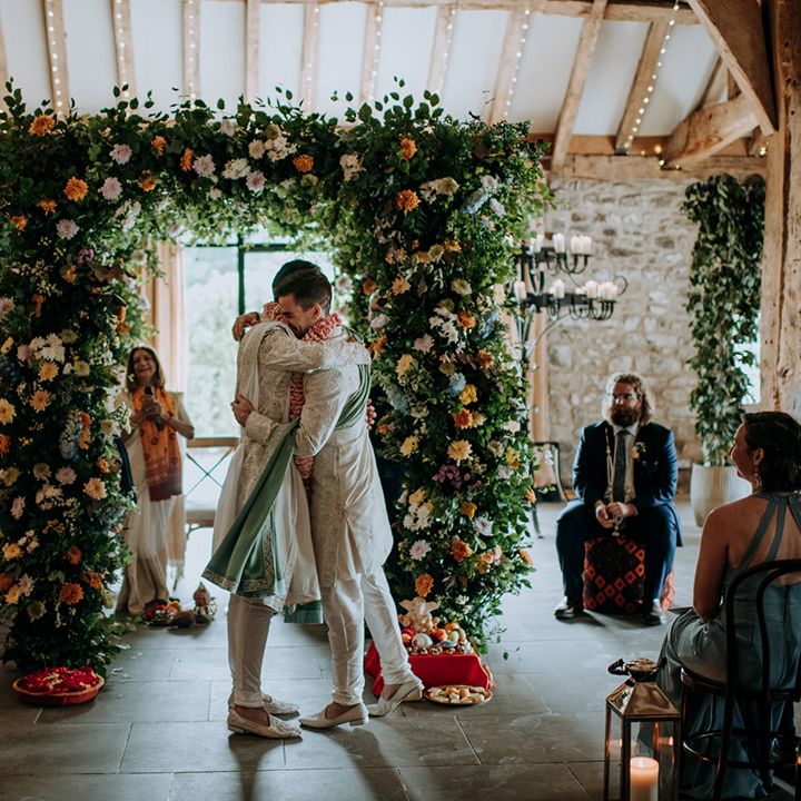 Grooms embrace beneath floral arch complete with blue and orange flowers as lights hang from the beams above
