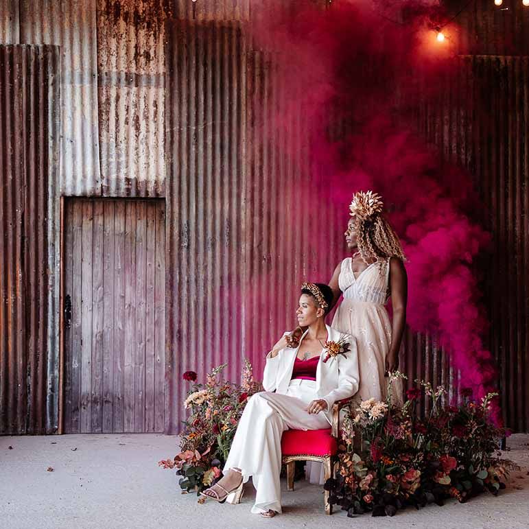 Bride in sleeveless floaty peach gown with embroidered stairs and gold leaf crown and bride in elegant white silk suit with autumnal dried floral boutonniere sitting on gold and red velvet chair with deep pink smoke bomb and dried autumnal arrangement