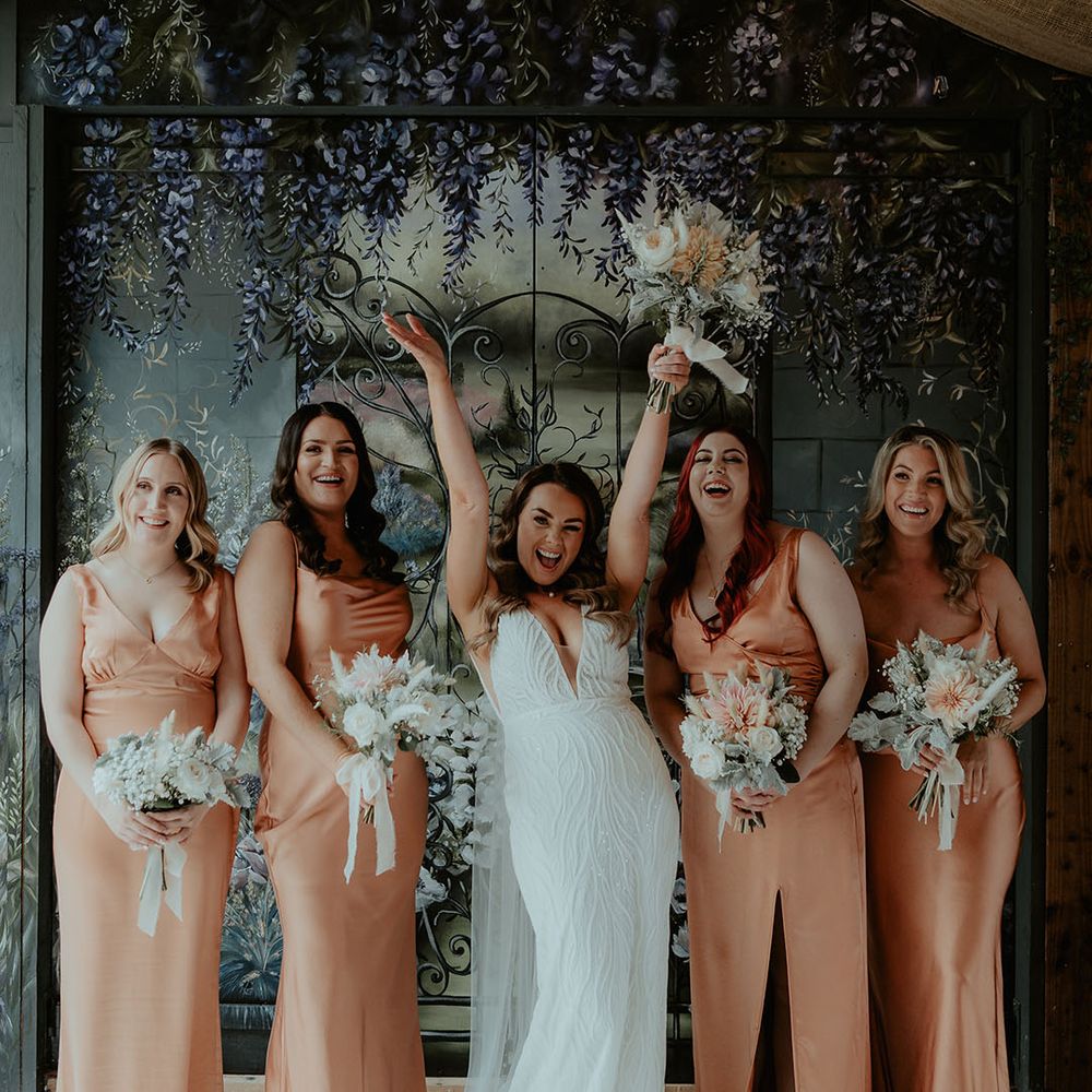 Bridesmaids in peach satin bridesmaid dresses holding white rose and gypsophila bouquets with bunny grass with the bride for the bridal party photos 