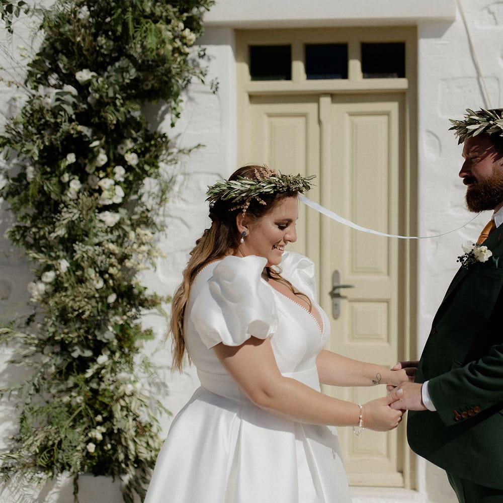 Bride and groom wearing green floral crowns and holding hand at intimate wedding ceremony with green and white flower column decor