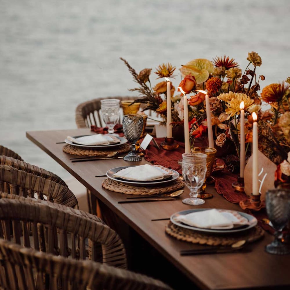 Rustic wedding tablescape by the Mexican sea with wicker chairs, burnt orange dried roses, dried flower and foliage floral centrepieces, wooden slate wedding table numbers, light orange tapered candles and burnt orange table runners