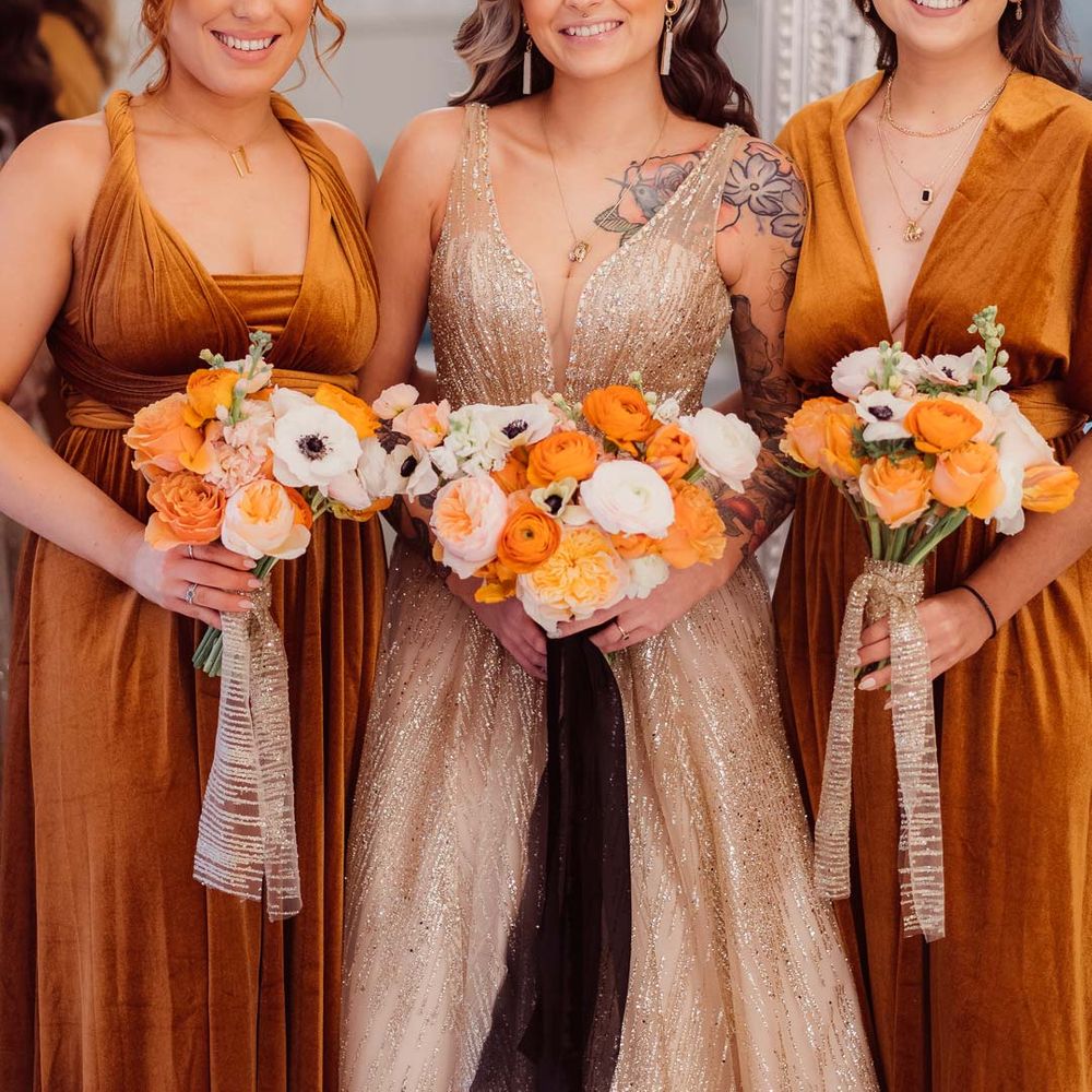 Bride in sleeveless deep v neck sparkly gold wedding dress with sheer and gold polka dot wedding veil standing with bridesmaids in velvet burnt orange bridesmaid dresses all holding orange garden rose, white poppy, light pink carnation and foliage bridal bouquets wrapped in sparkly gold ribbon