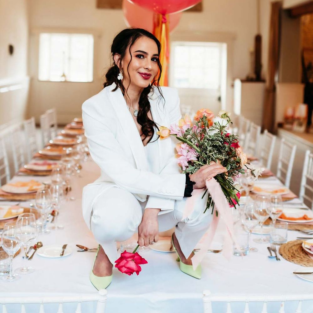 Bride white bridal suit with silver bridal jewellery with closed toe bright green wedding heels squatting on wedding tablescape with white wedding tablecloth, burlap place mats, tapered candles, pink and orange wedding stationery, and floral wedding table centrepieces with pink and orange garden roses, orange floribunda, yellow ranunculus flowers, foliage and Iceland poppies 