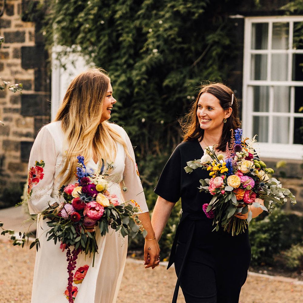 Two brides at same sex wedding with bride wearing colourful embroidered wedding dress and bride in bridal black jumpsuit holding colourful bouquets