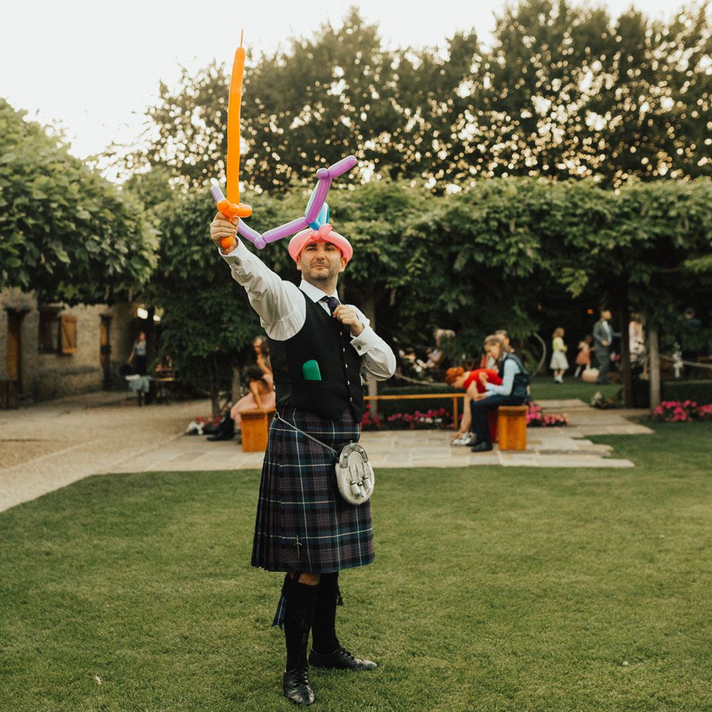 Wedding guest in kilt with inflatable sword poses outside at Tythe Barn Wedding with boho decor 