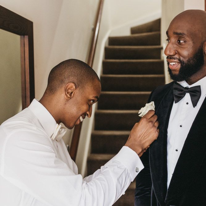 Groomsman pinning the Grooms white rose buttonhole onto his velvet tuxedo jacket 