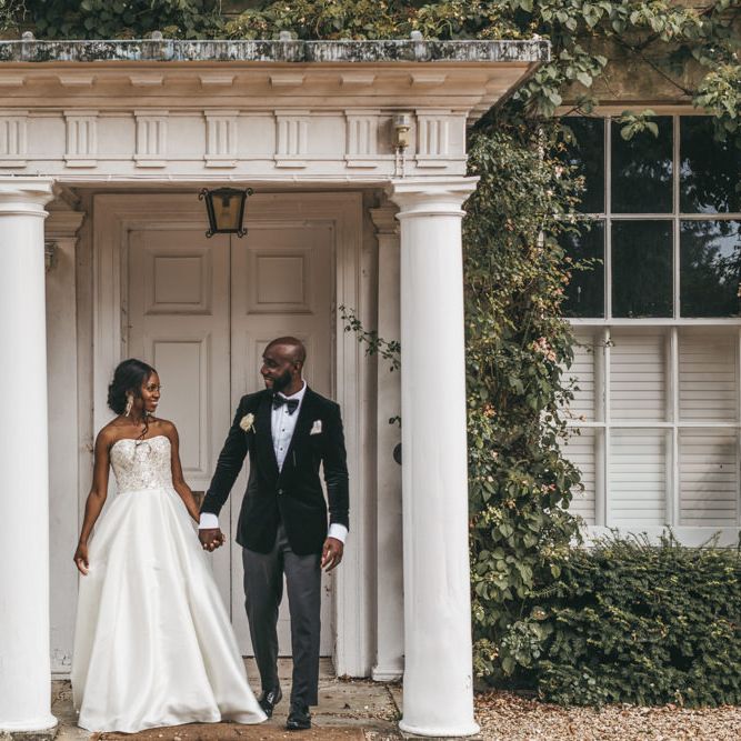 Black bride in a strapless Oleg Cassini wedding dress with embellished bodice holding her grooms hand in a velvet tuxedo outside Northbrook Park 