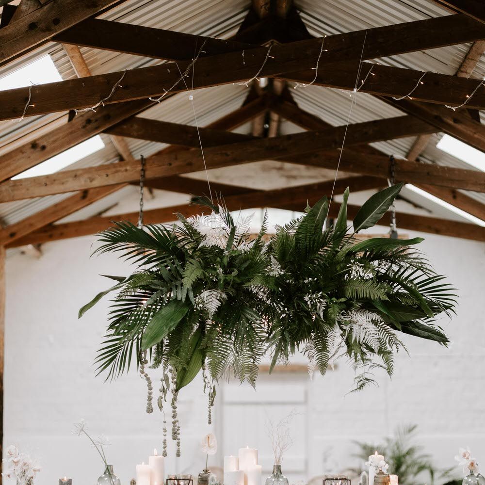 Tropical wedding table with industrial chic vibes and wooden bar stools, underneath a hanging installation of tropical leaves