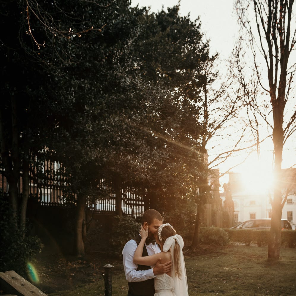 Golden hour portrait with the bride and groom kissing. The bride wears a long bow wedding veil 