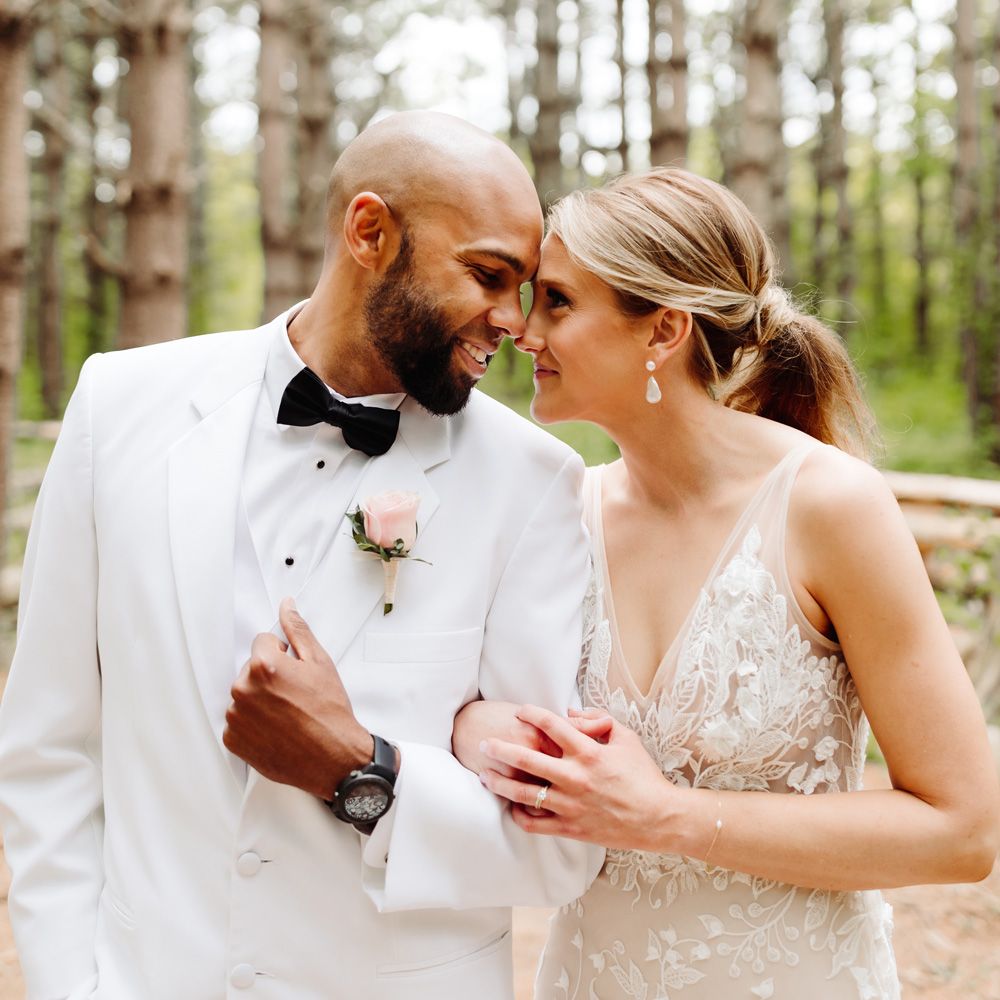 Groom in white tuxedo jacket and pink rose buttonhole links arms with bride in white Made With Love wedding dress as they touch foreheads in woodland after backyard wedding