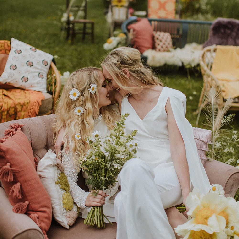 Bride in a jumpsuit with half up half down hair sitting on her brides lap on a retro sofa at outdoor wedding ceremony 