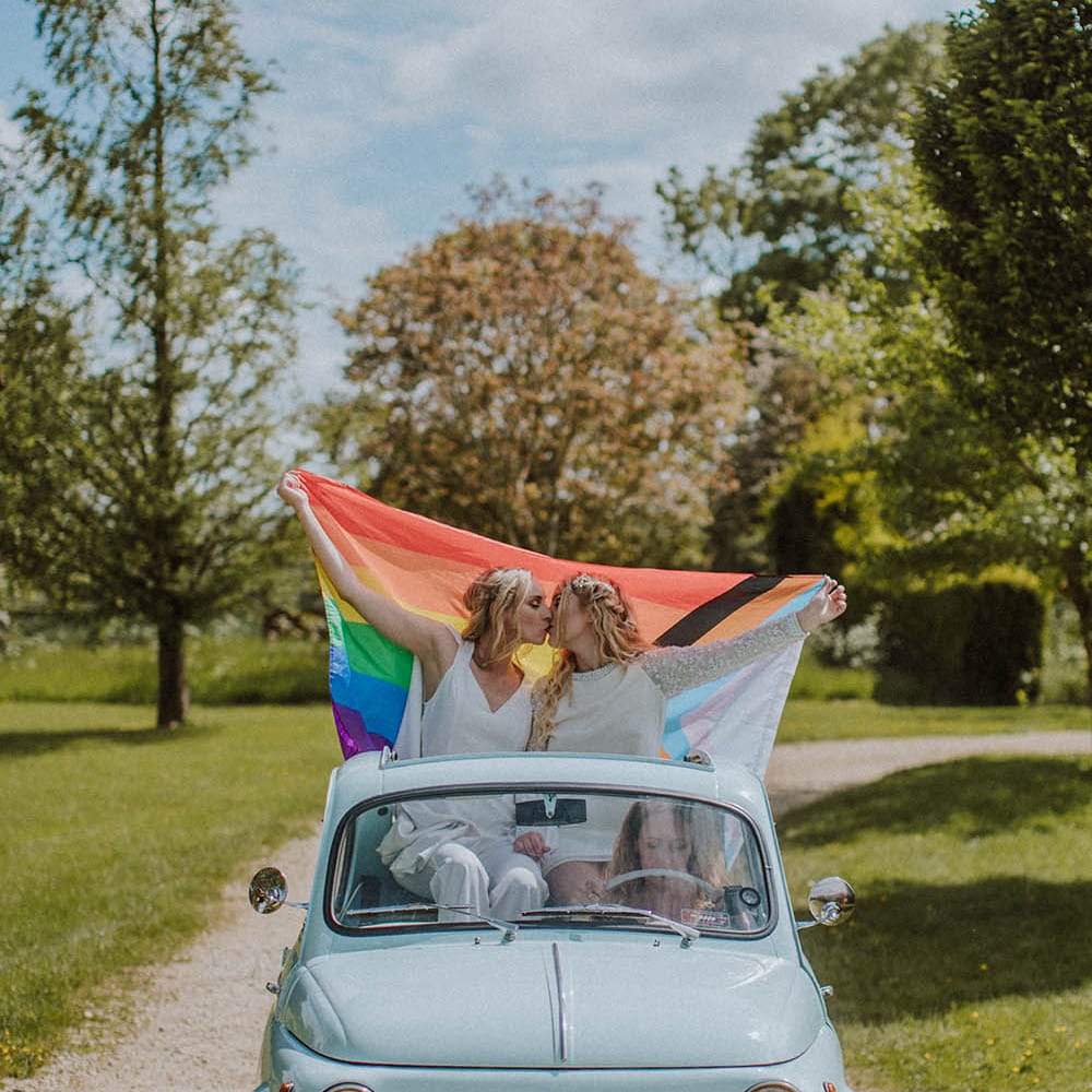 Two brides kissing and holding a Pride flag on the back of the light blue fiat 500 car 