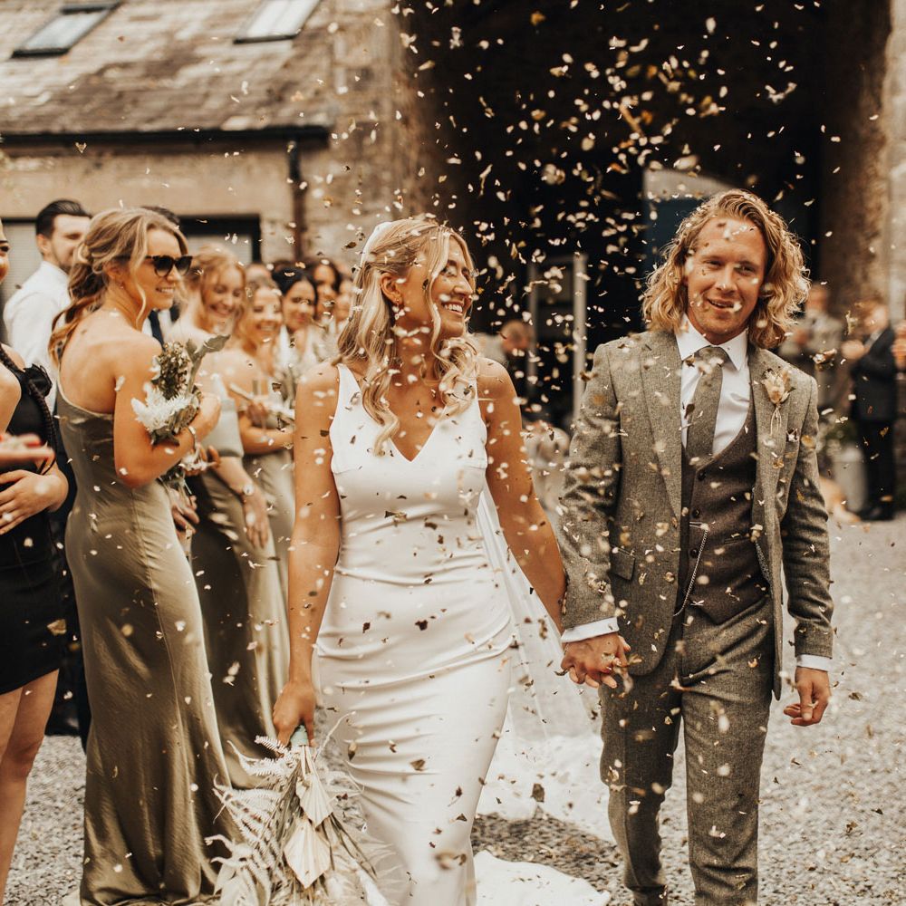 Smiling bride in white V neck Made With Love wedding dress holding dried bridal bouquet holds hands with groom in sage green suit as they walk through courtyard of guests throwing confetti at Anran Devon
