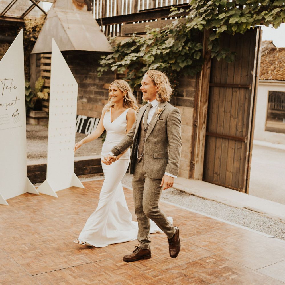 Bride in white Made With Love wedding dress holds hands with groom in sage green suit as they walk into glasshouse wedding breakfast at Anran House