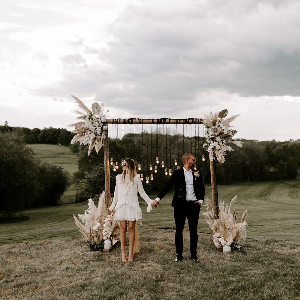 Stylish bride in a short wedding dress and sunglasses holding hands with her groom in a black suit in front of a wooden frame altar decorated with lights and dried flowers 