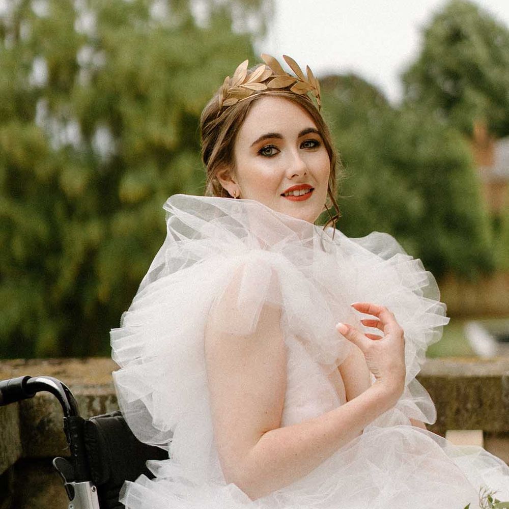 Bride in a tulle wedding dress and gold crown sitting on the balcony at Bourton Hall in her wheelchair with Crocheted wheelchair covers