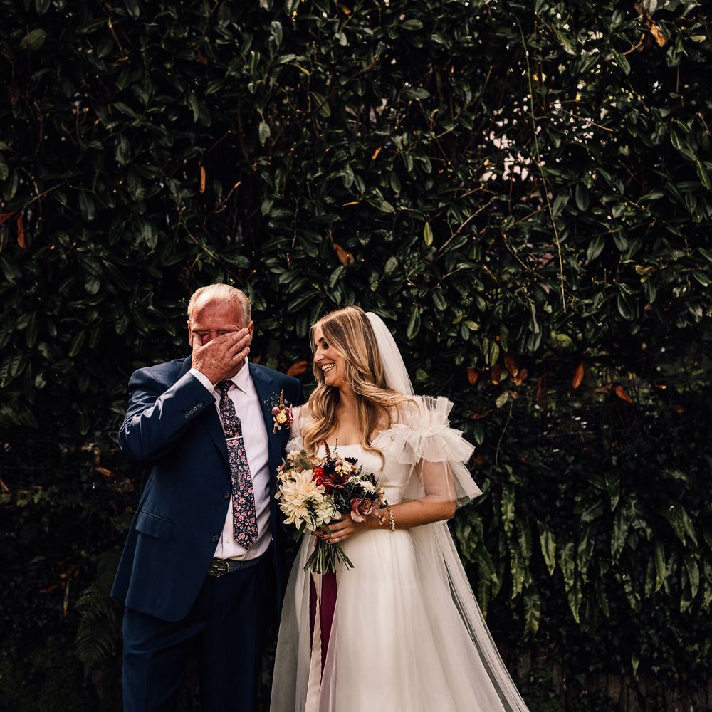 Bride in white Halfpenny London Mayfair dress stands outside holding mixed bridal bouquet with burgundy ribbon with emotional father in blue suit before church wedding ceremony in Cornwall