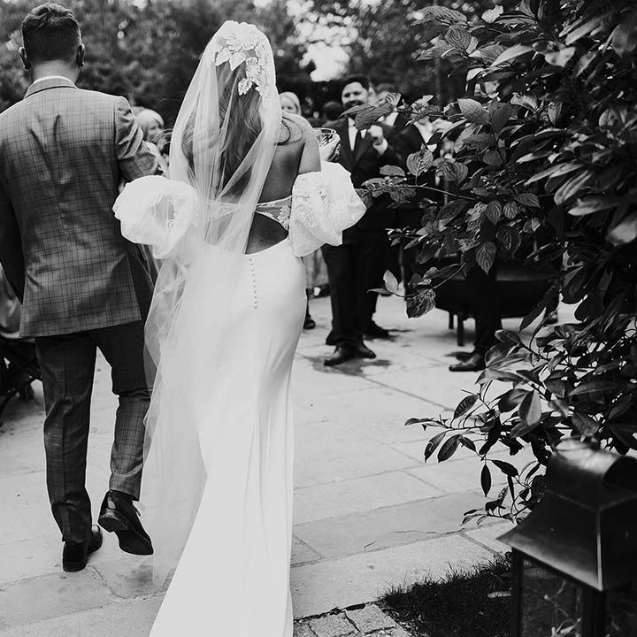 Bride & groom walk together in black & white image as bride wears lace embellished veil 