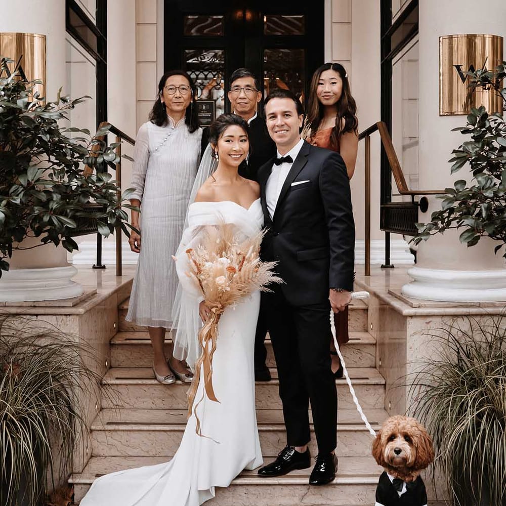 Bride in simple cross over wedding dress smiling with groom outside Chelsea Town Hall with the family wedding party and a cockapoo dog