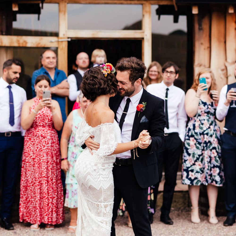 Bride in off the shoulder Grace Loves Lace wedding dress, dusty pink block heels and colourful flower crown dances with groom in black suit and colourful floral buttonhole outside The Bridal Barn wedding venue as wedding guests watch