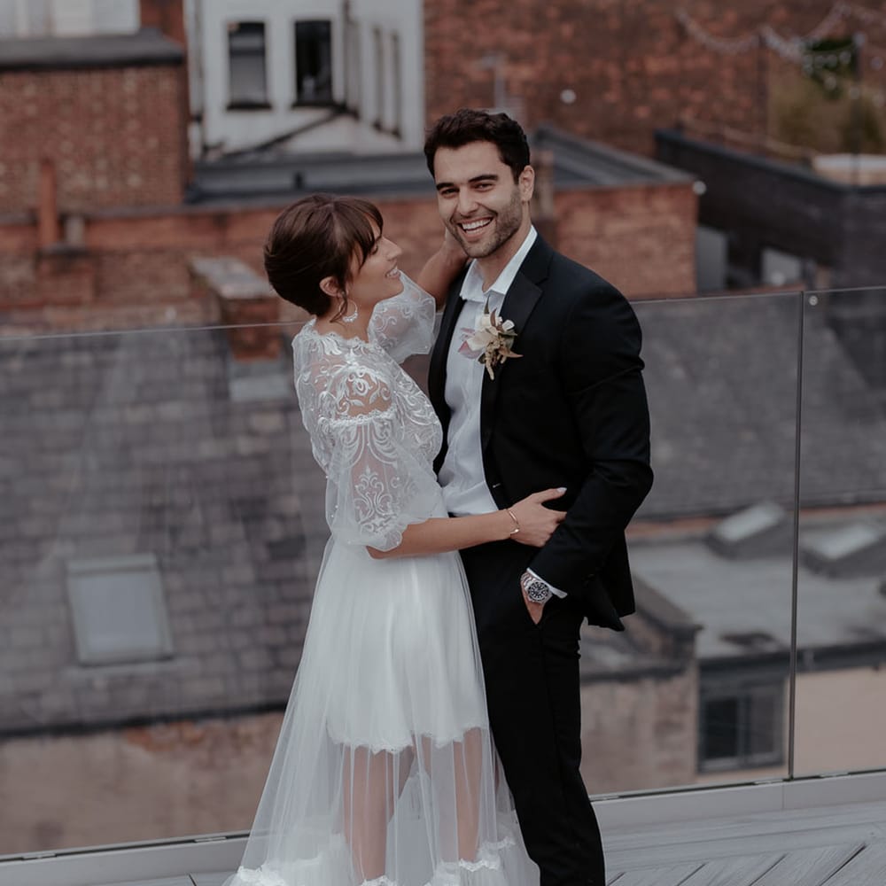 Bride in a short wedding dress with lace bodice and floor length tulle over skirt embracing her groom in a black tuxedo 