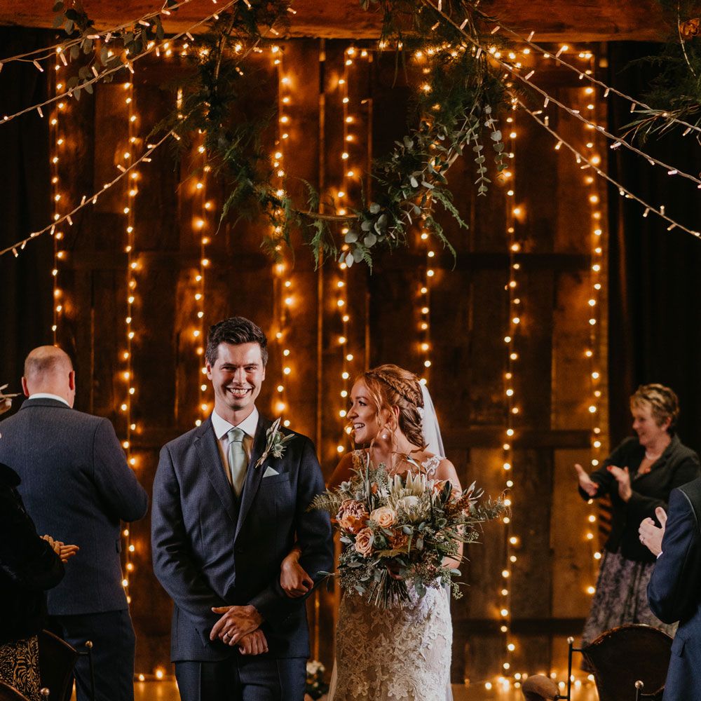 Bride in lace and tulle wedding dress holding large mixed bridal bouquet walks down the aisle with groom in dark suit and light blue tie for fairy lit wedding ceremony