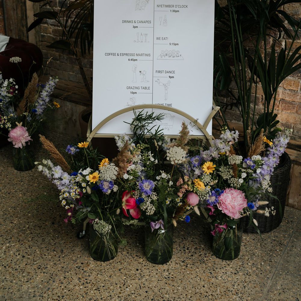 Order of the day wedding sign with mini cartoon style sketches and colourful flowers in glass vases