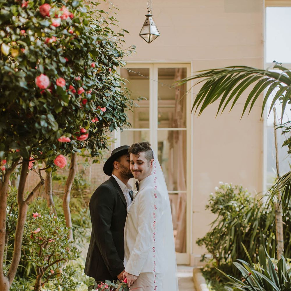 Groom in a black suit, fedora hat and boots kissing his partner in a white groom suit and patterned edge veil at Prestwold Hall Barns
