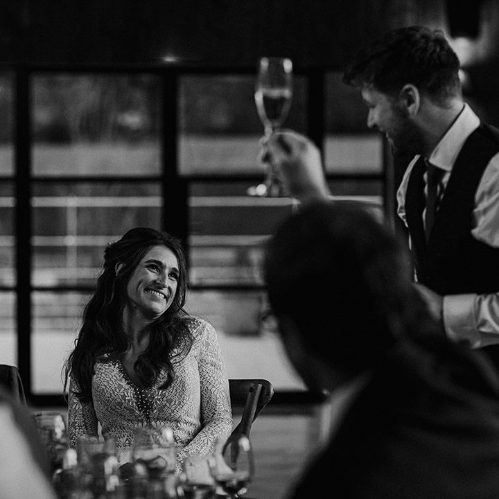 Bride in illusion lace wedding dress smiles adoringly at the groom giving his speech 