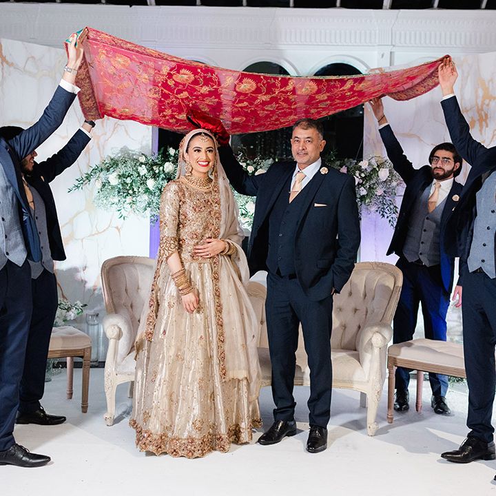 Bride & groom stand beneath brightly coloured fabric held up by wedding guests after Nikkah & Walima