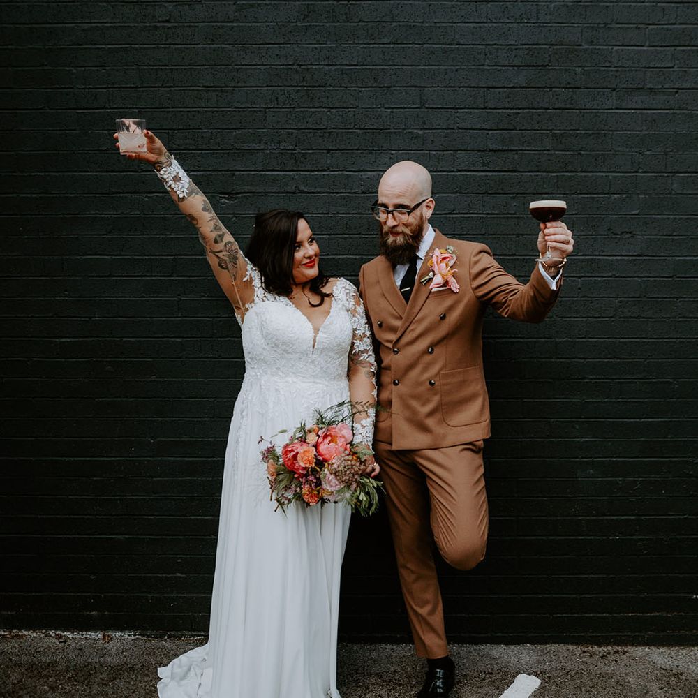 Bride wears lace wedding dress with sheer sleeves whilst stood beside her groom in brown suit with statement pink floral buttonhole