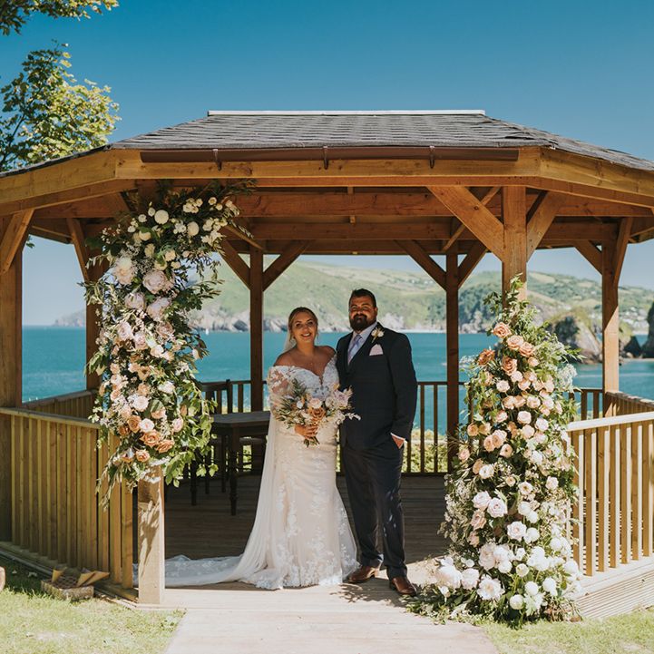 Bride & groom stand beneath wooden pergola surrounded by floral installation with green foliage 