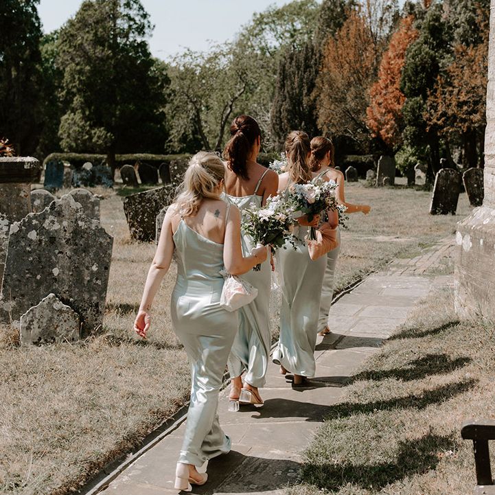 Bridesmaids walk in a row in satin sage green dresses with bouquets to the church for the ceremony 