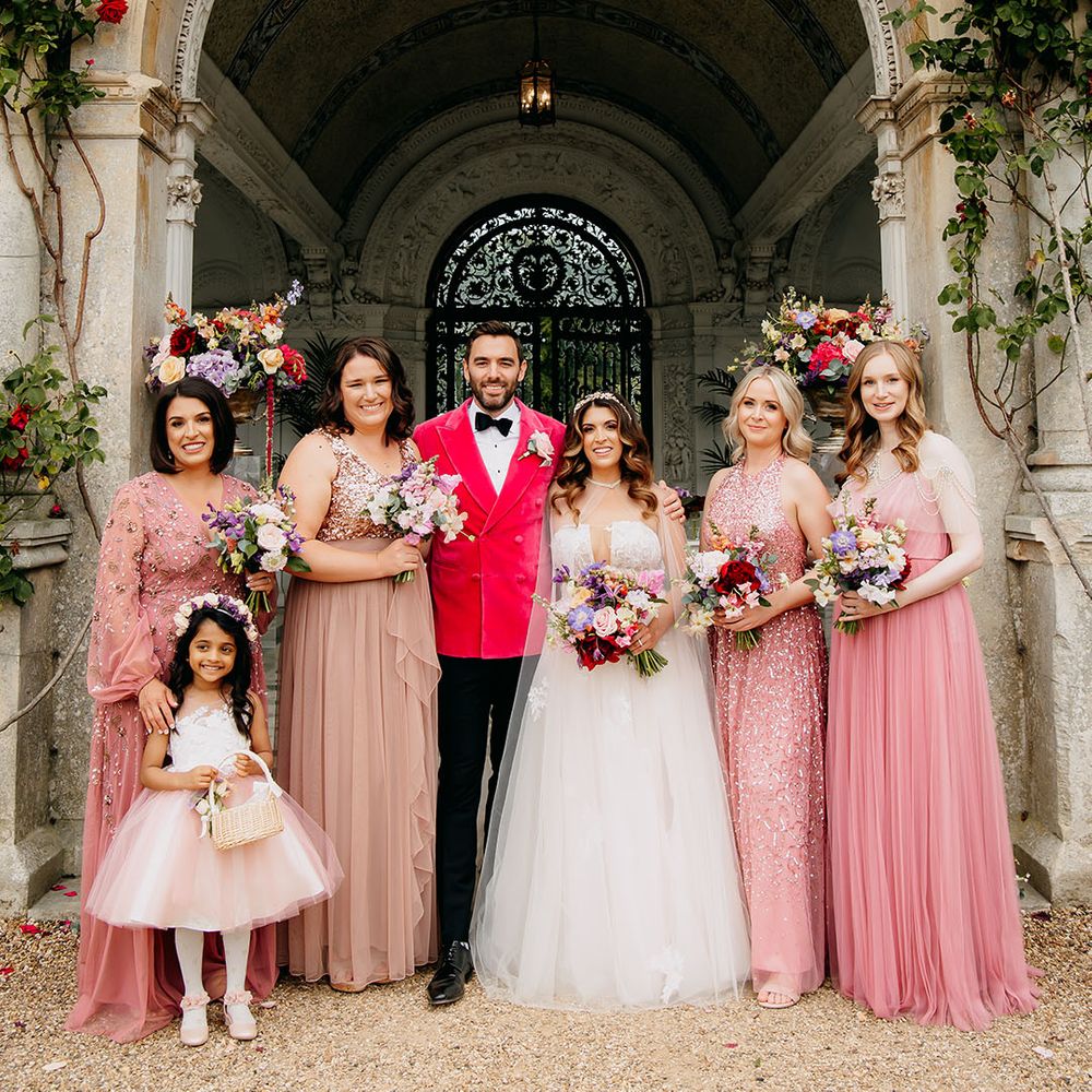 Bridesmaids in mismatched pink sparkly dresses with flower girl in a flower crown and short pink tulle dress with the bride in a sheer cape and groom in bright pink velvet jacket