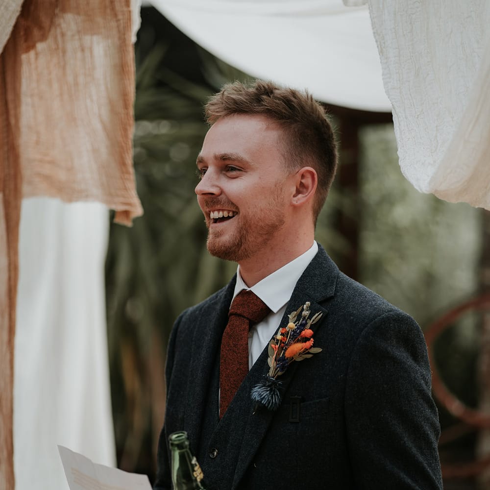 Groomsmen in dark grey suit, burgundy tie and dried flower boutonniere doing wedding speech surrounded by burnt orange and white wedding drapes decorations 