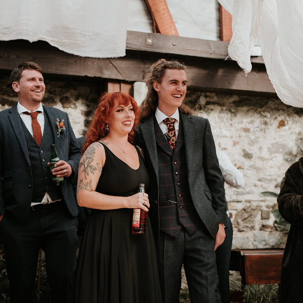 Bride in strappy black dress for wedding in Devon, standing with groom in dark grey three piece suit and burgundy flower patterned tie listening to wedding speeches 