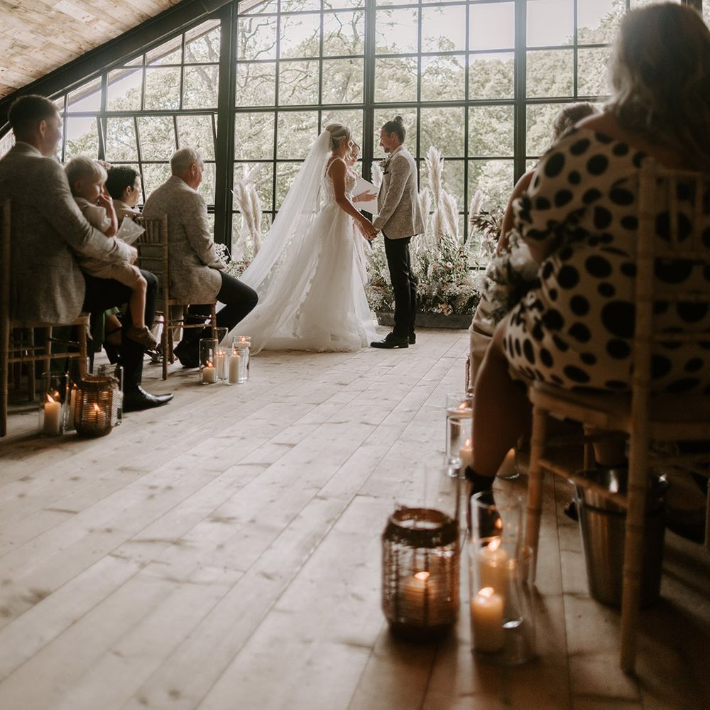 Hidden River Cabins wedding venue in Cumbria with big windows decorated with candles in vases along the aisle and pampas grass at the altar