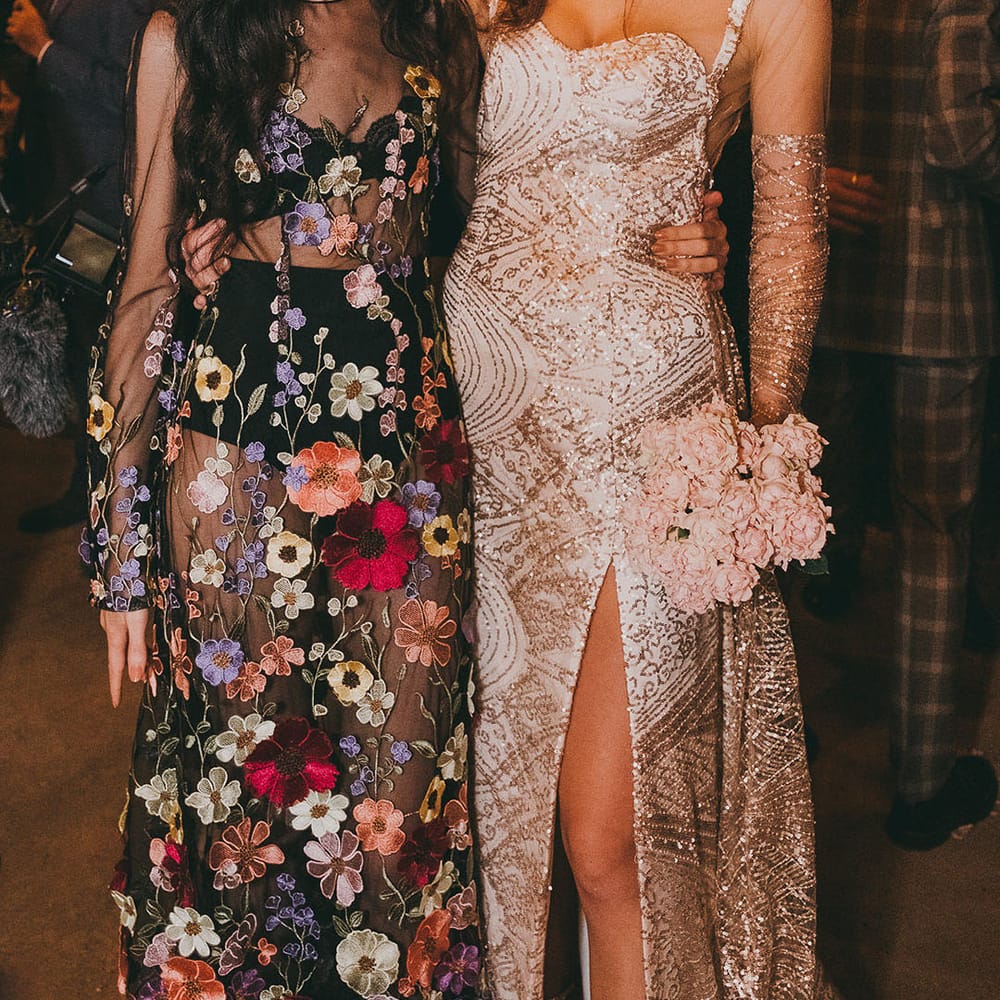 Bride in a bespoke gold sparkly wedding dress with illusion sleeves embracing her wedding guest in a flower appliqué dress