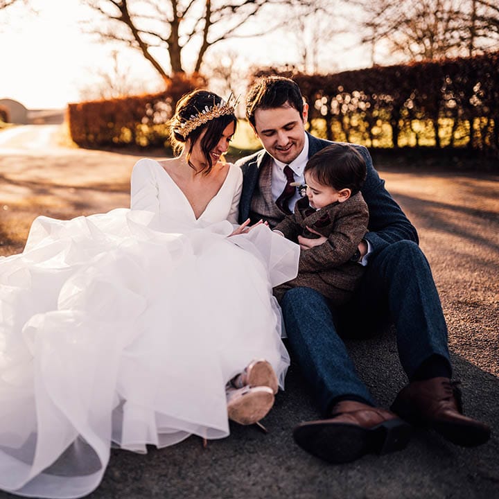 Bride in ruffle gown wearing bride crown sitting with groom in navy suit and their little boy in brown suit 
