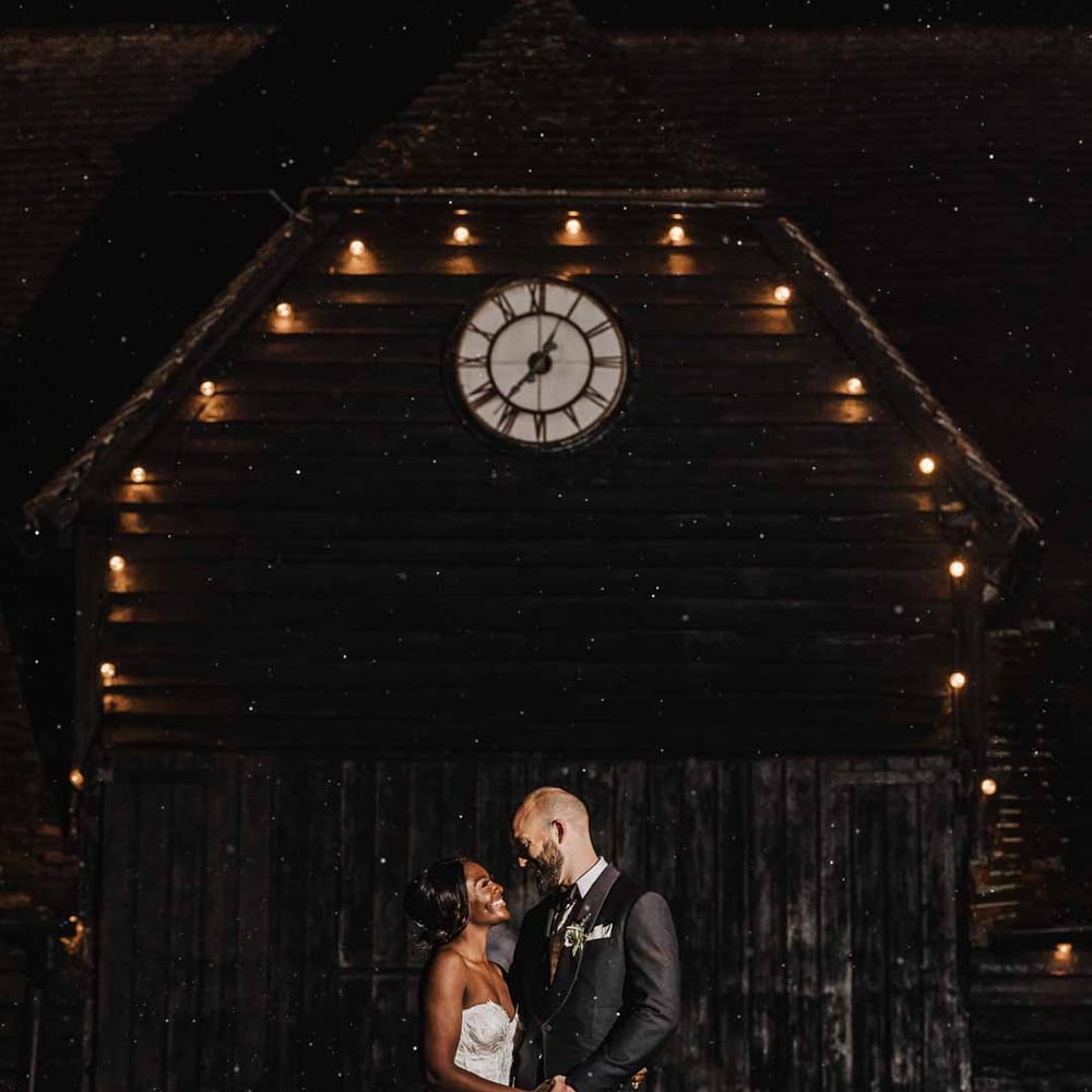 Groom in classic black three piece grooms suit with bow tie and white rose and foliage boutonniere in church standing with bride in sweetheart neckline lace wedding dress outside of Lains Barn 